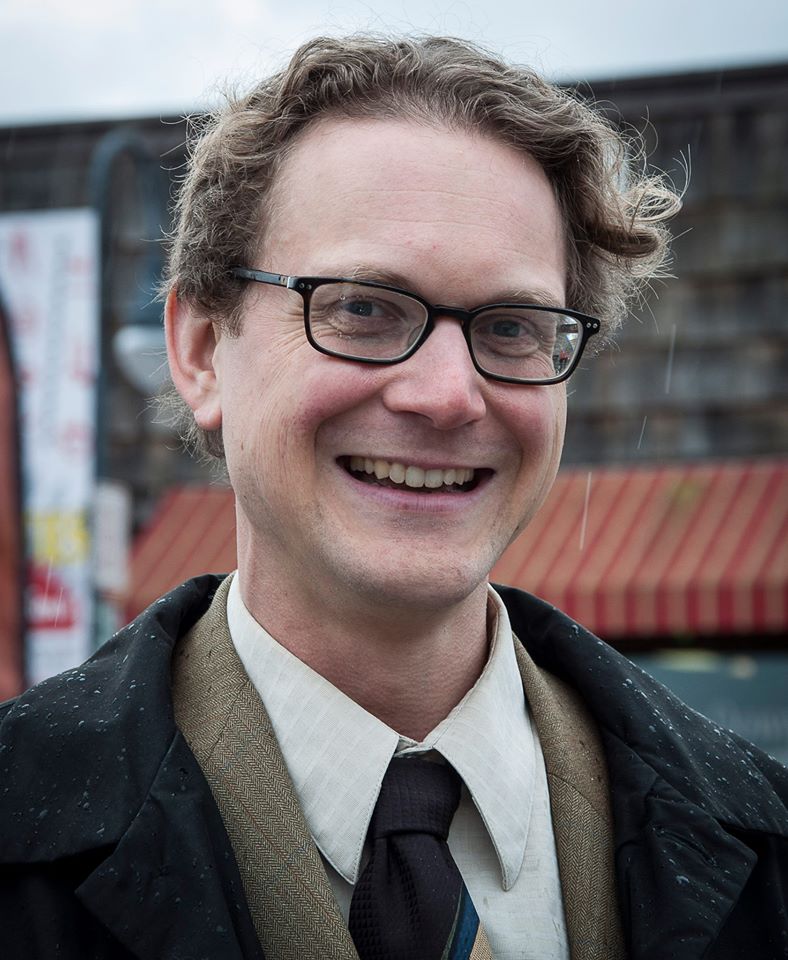 Photo of Lindsey Schromen-Wawrin who has short curly brown hair, is wearing black framed glasses, a brown suit, black coat and tie.