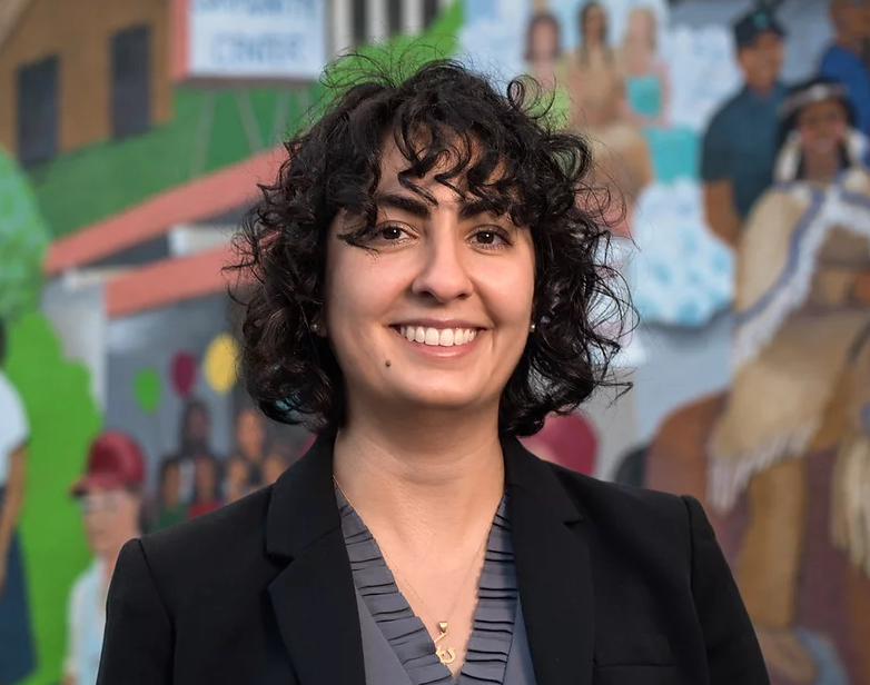 Headshot of Darya in front of a mural. She has black curly hair, is smiling, and wears a suit jacket