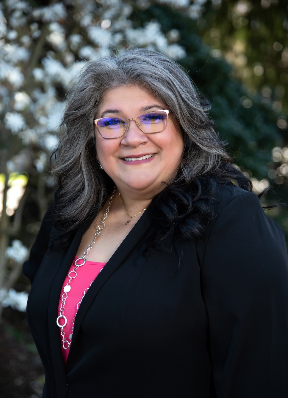 Photo of Diana Perez wearing a dark pink blouse and a black blazer. She has long black and grey wavy hair.