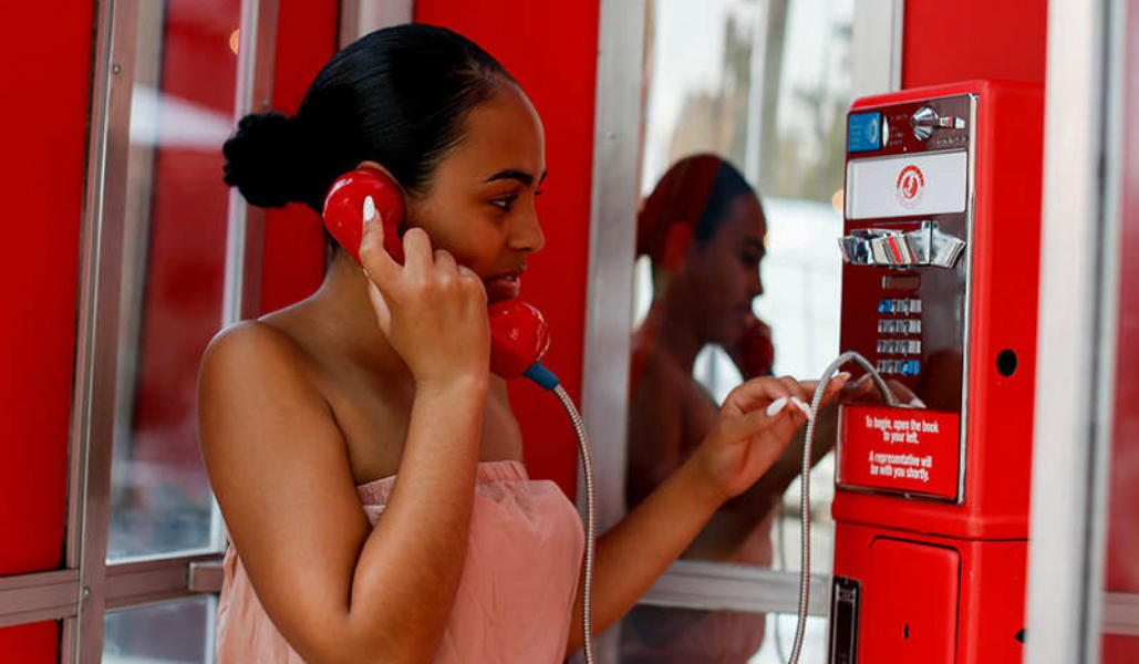 Photo of woman using a red telephone booth. She has dark hair in a bun and a peach blouse.