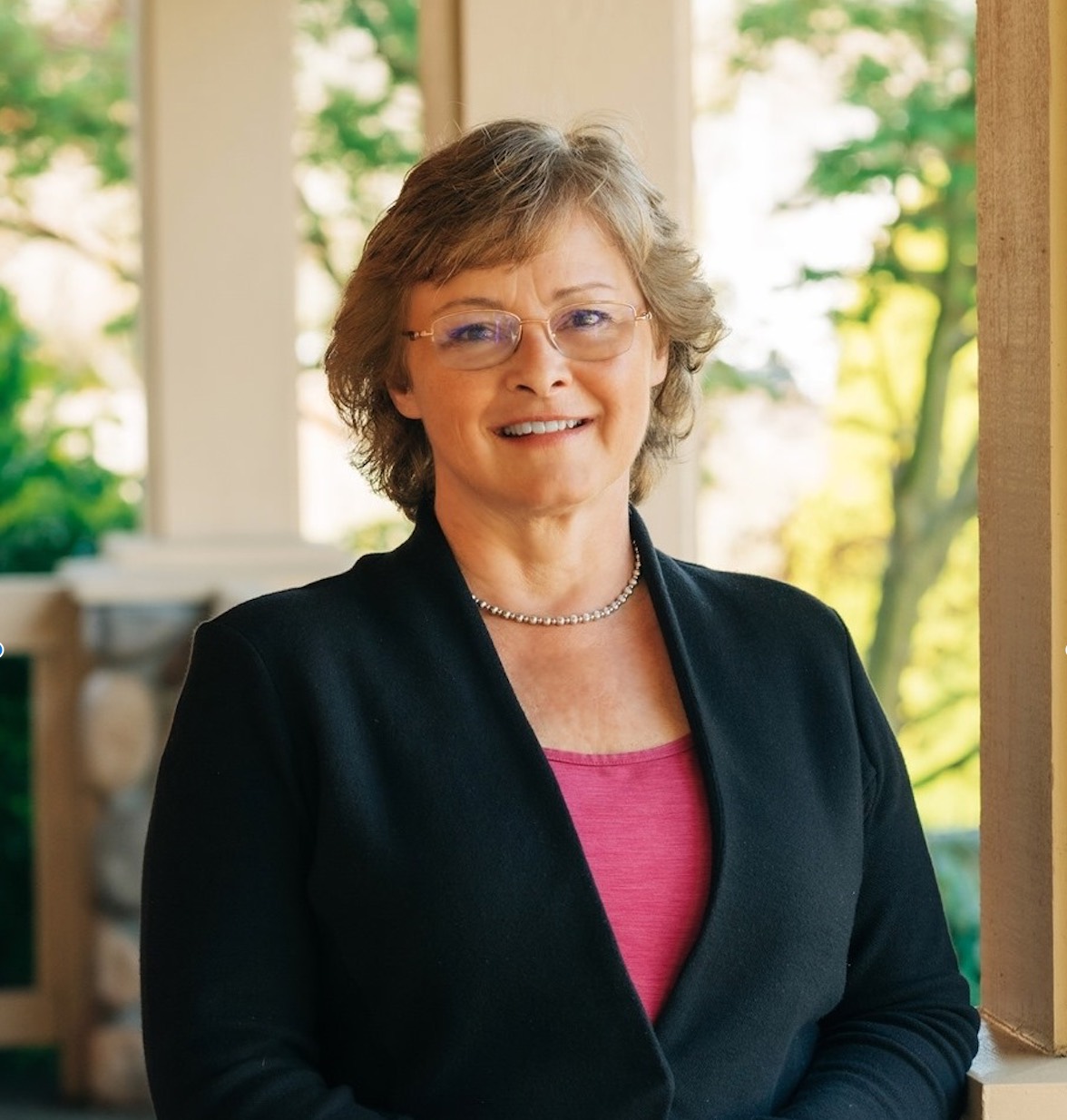 Photo of Sarah Perry who has wavy light brown, short hair. She is wearing glasses, a bright pink top and black blazer.